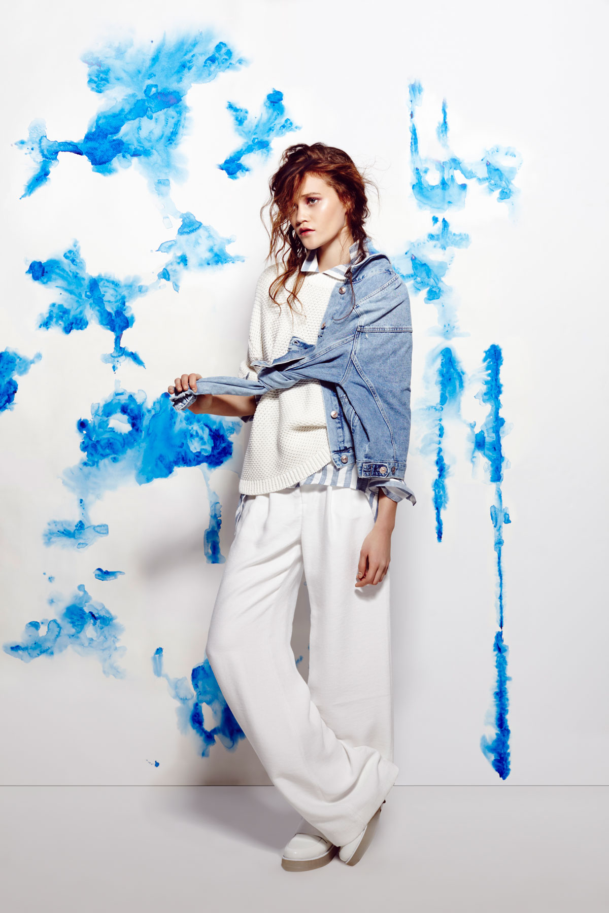 Creative fashion campaign featuring a model in a white knit outfit and denim jacket against a vivid blue painted backdrop.